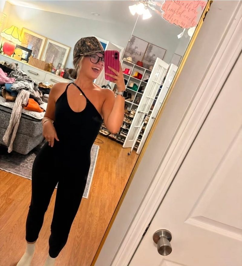 Client in a black jumpsuit and cap, smiling in a mirror photo with a natural-looking full-body glow