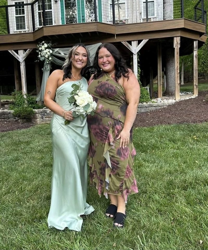 Two clients at an outdoor wedding, one in a sage satin gown holding a white bouquet, both with a natural-looking tan