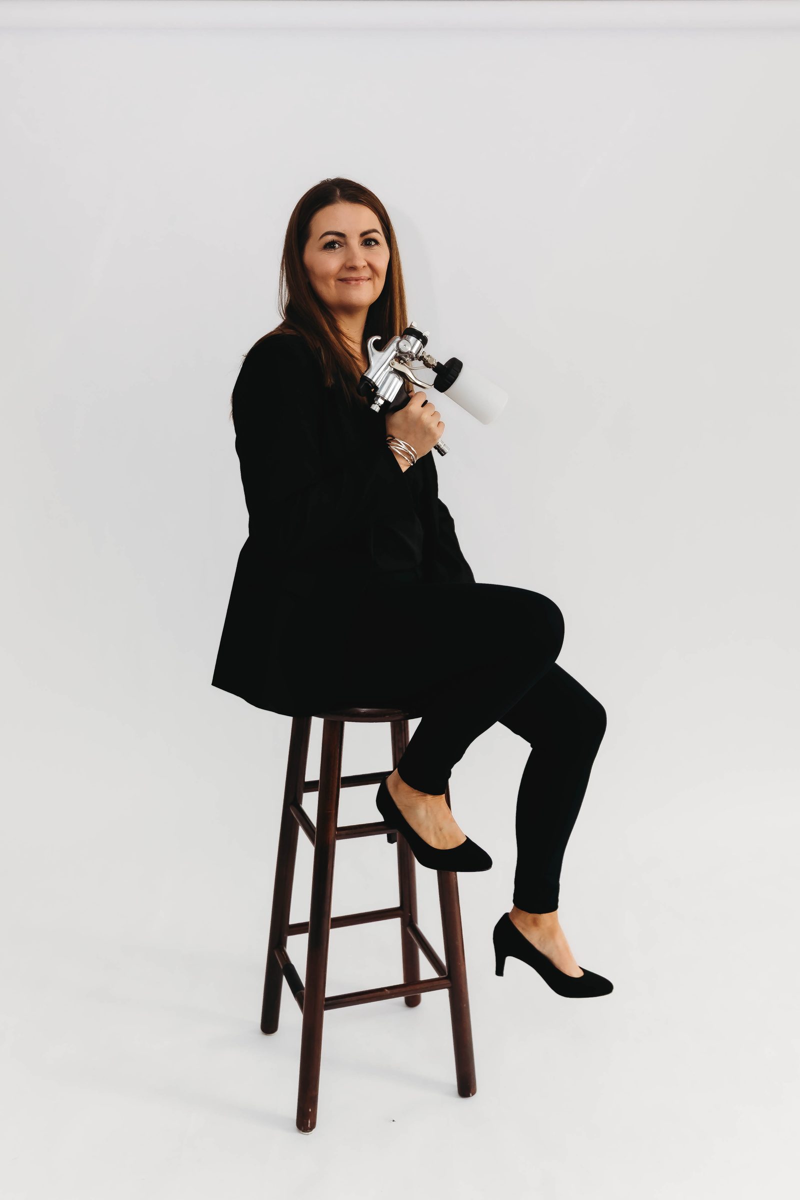 Seana, owner of The Sunless Method, seated on a stool in her Brentwood studio holding her airbrush gun