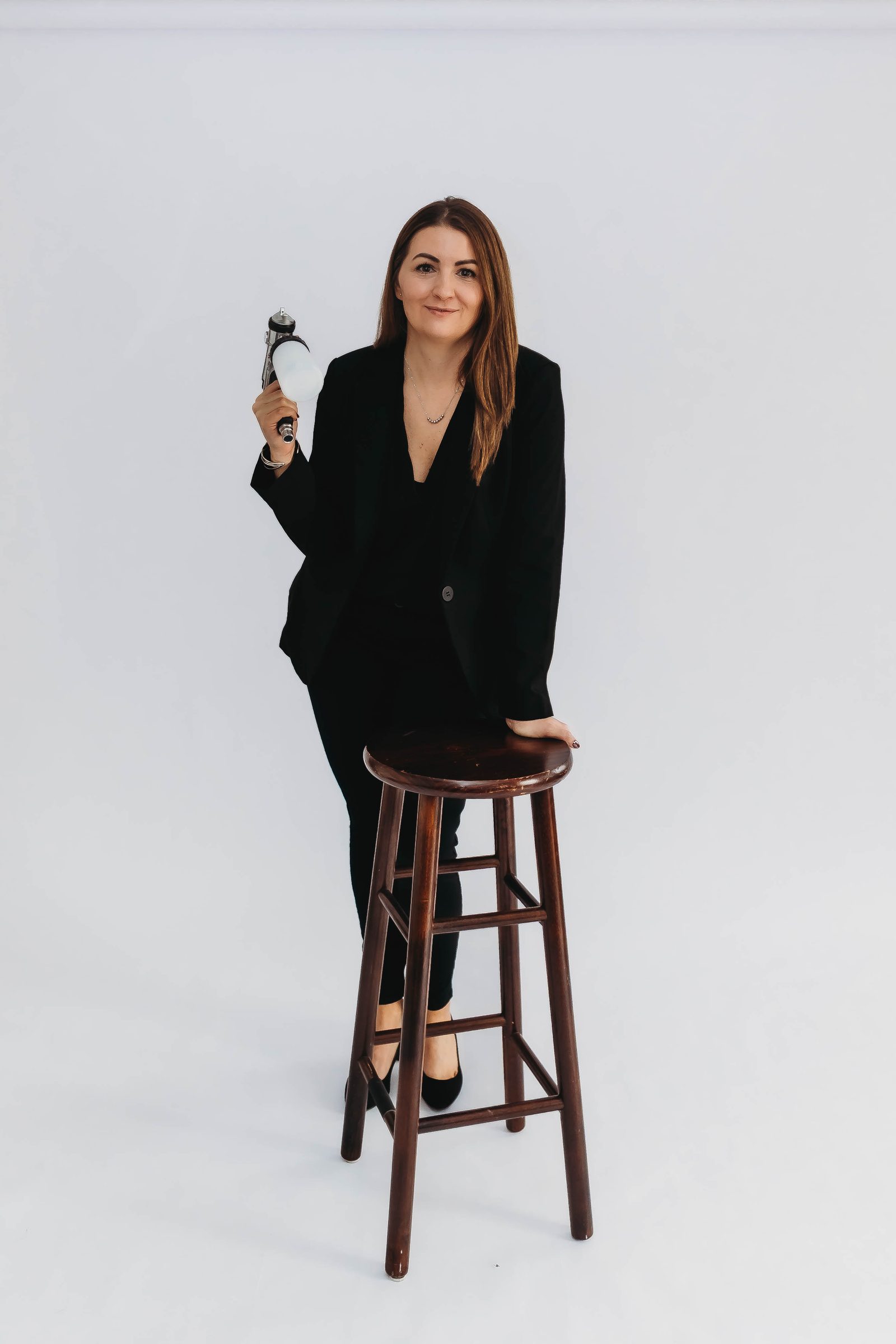Seana standing beside a wooden stool in a black suit, holding her airbrush gun