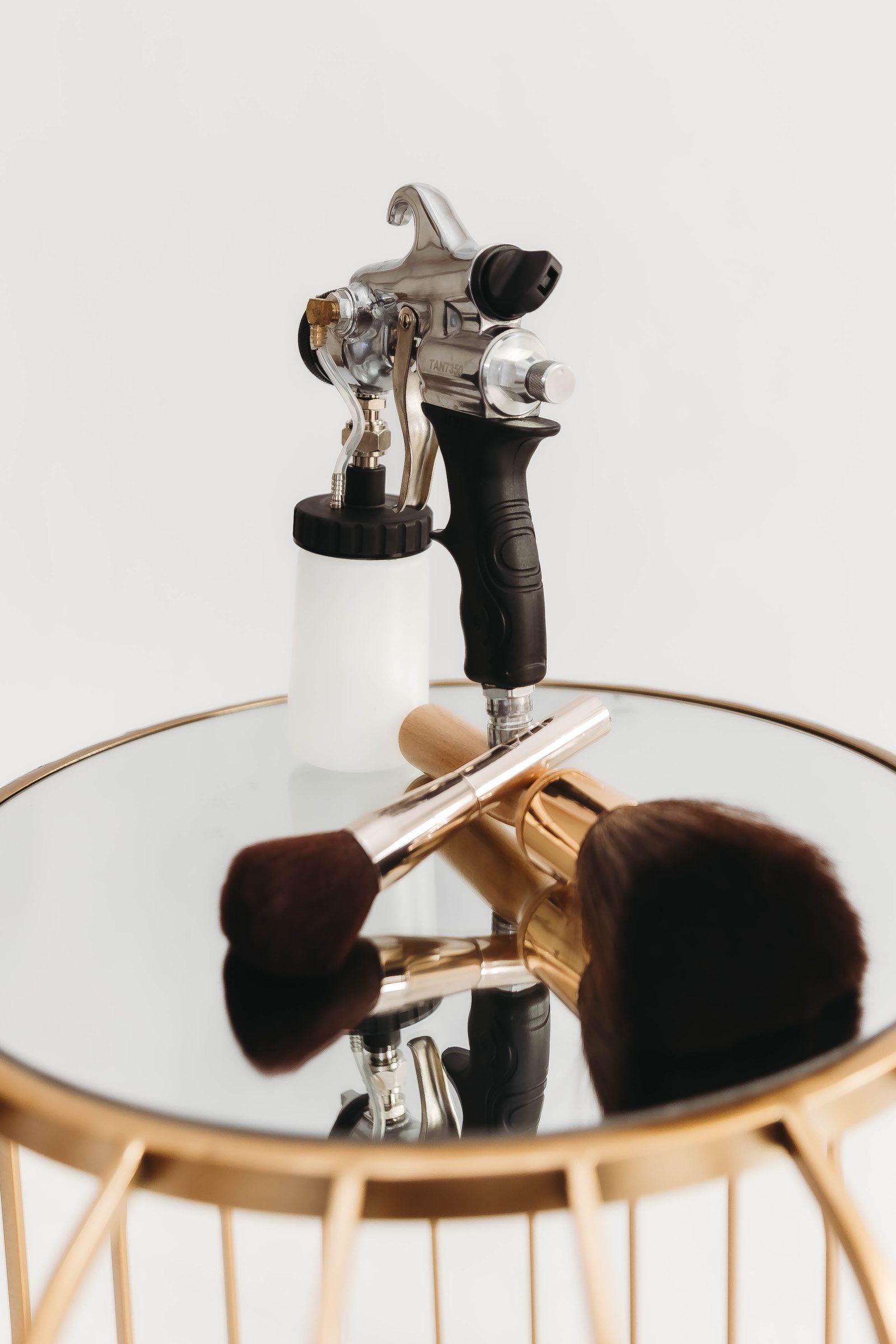 A professional airbrush spray tan gun and two finishing brushes on a mirrored gold side table in the studio